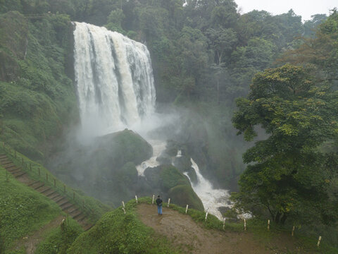 Aerial view of a majestic waterfall cascading down mossy rocks, surrounded by lush greenery, creating a misty spectacle, Curugsewu, Central Java, Indonesia.