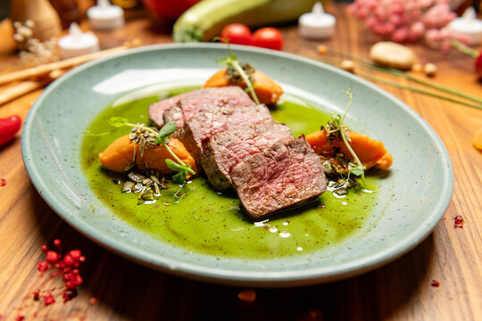 Juicy beef steak slices on rich green sauce with carrot puree and microgreens, beautifully plated on ceramic plate, rustic wooden table