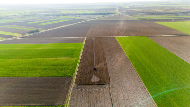 Aerial view of vibrant green and brown fields, divided by straight lines, where a single tractor tills the soil, Sremska Mitrovica, Vojvodina, Serbia.