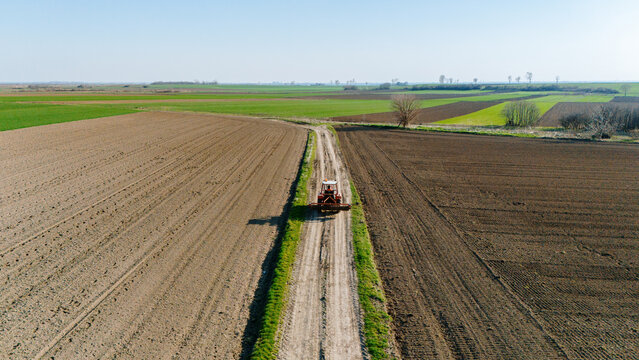 Aerial view of a lone red tractor kicking up dust on a pale dirt road slicing through fields of tilled brown earth and vibrant green crops, Sremska Mitrovica, Vojvodina, Serbia.