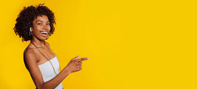 Happy young black woman with afro hair smiling and pointing finger on yellow background.