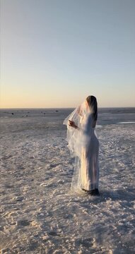 Elegant woman in veil standing on frozen sea at sunset