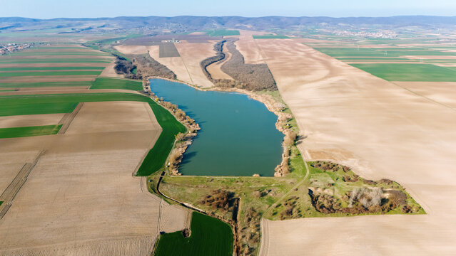 Aerial view of an idyllic lake reflecting the clear sky, surrounded by a patchwork of golden and green fields, Mandelos, Vojvodina, Serbia.