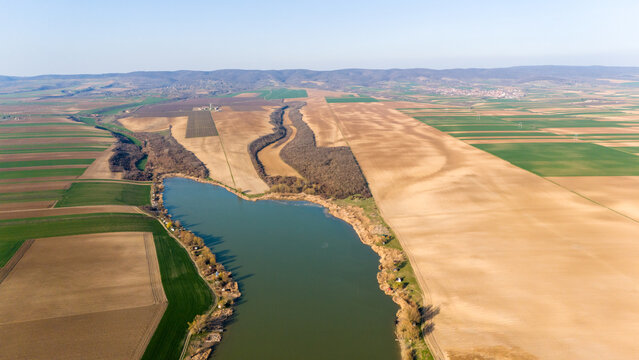Aerial view of a serene lake reflecting the sky, bordered by fields of contrasting greens and browns, framed by distant hills, Mandelos, Vojvodina, Serbia.