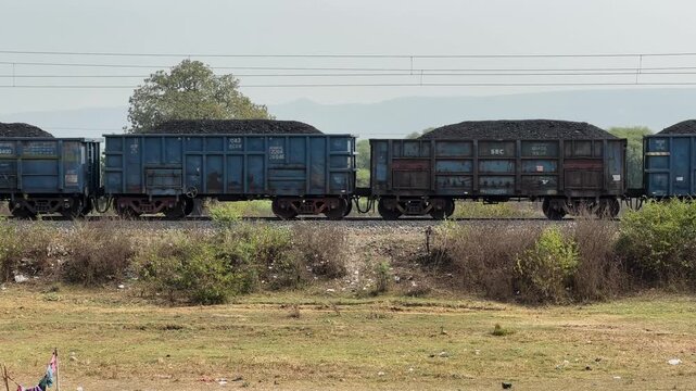 Side view of freight train wagons loaded with coal moving along a railway track through a rural landscape in India, showing bulk cargo transportation for energy and industry.