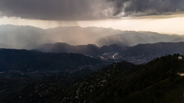 Aerial view of rain cascading over the rugged, forested mountains, casting shadows on the valleys below, Pir Chanasi, Azad Kashmir, Pakistan.