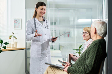 Obraz premium Young female doctor smiling while talking to senior man and senior woman sitting in medical office waiting area, holding clipboard and digital tablet