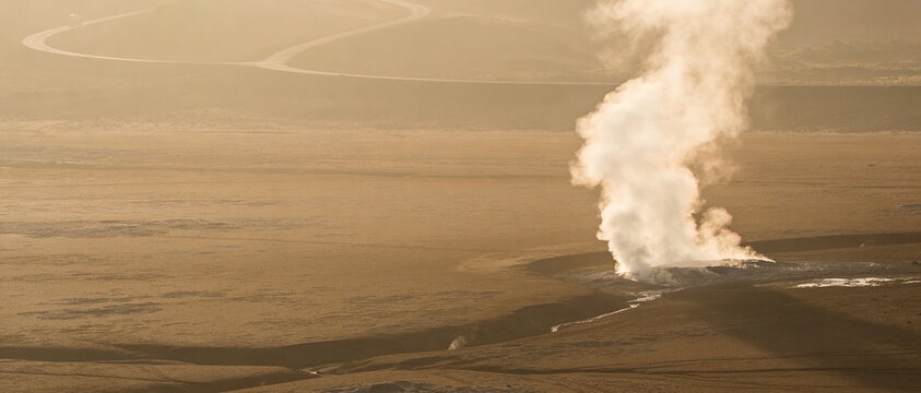 Wide shot of geothermal vent with rising steam against desert landscape and winding road.
