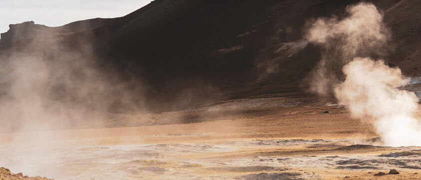 Steam rising from hot springs against brown volcanic mountains in sunny day.