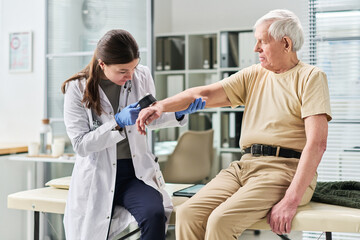 Obraz premium Female young doctor examining arm of senior man using dermatoscope in medical office, both focused on procedure, senior man sitting on examination table