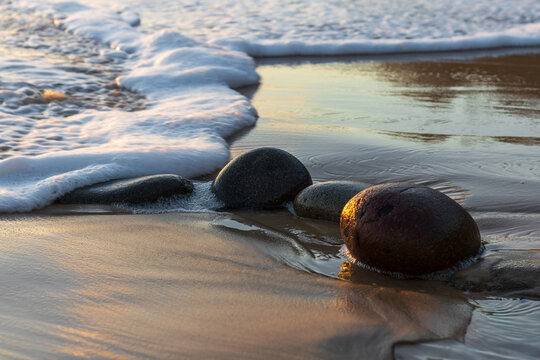 Ocean wave with sea foam washing over smooth round rocks on wet sand