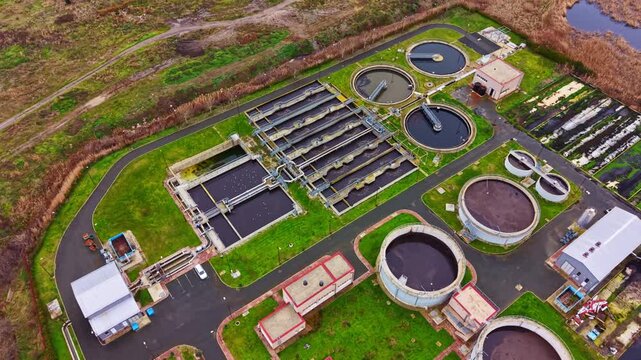 Aerial view shows a water treatment facility with multiple tanks and processing areas along with grassy space. The location is near a large water source and surrounded by nature.