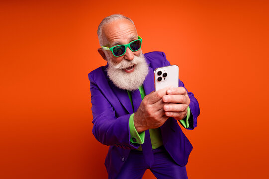 Elderly bearded man in a purple suit and green shirt selfies with a phone against a bold orange background showing retro style and joyful lifestyle
