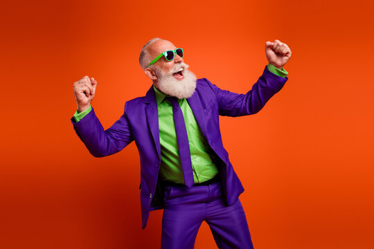Energetic elderly man in a vibrant purple suit dances with joy against an orange background wearing a green shirt and sunglass