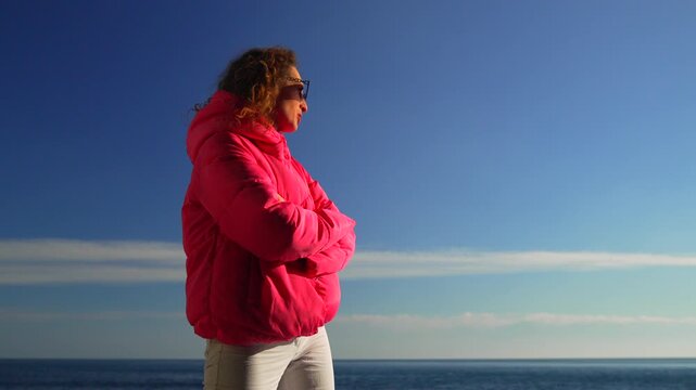Woman ocean jacket wearing a bright pink puffer standing by the sea under a clear blue sky on a sunny day