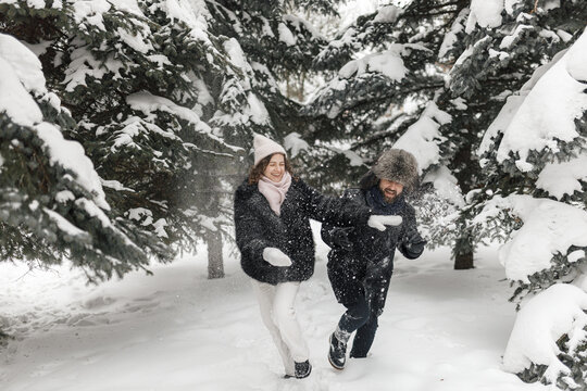 Cheerful active couple in fur coats running in snowy weather in winter forest and having fun 