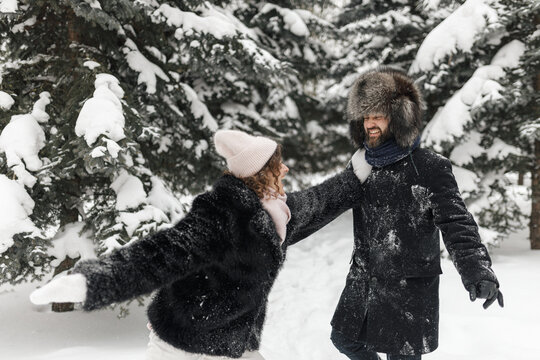 Happy playful couple in warm coats playing and having fun in winter forest among snowy trees 