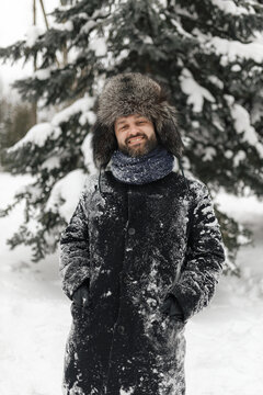 Portrait of bearded man with toothy smile wearing fur coat and hat looking at camera in snowy winter park 