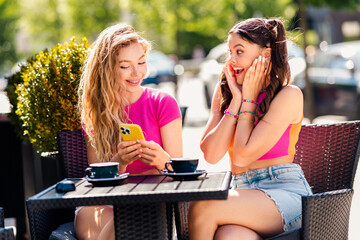 Two stylish young women enjoy a sunny urban cafe chat with phones in hand and bright smiles sharing laughter and friendship during a vibrant summer day in the city