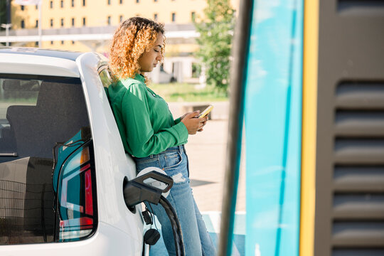 Woman leaning on electric car using smartphone at charging station outdoors