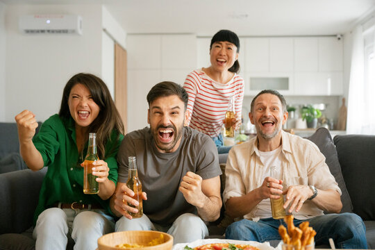 Friends cheering while watching football match at home with drinks