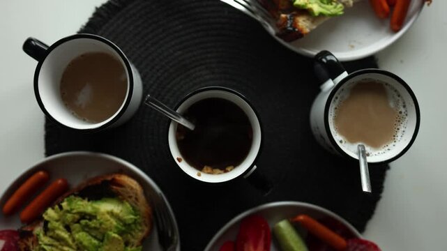 A table shows several mugs with different types of drinks. Plates hold slices of bread, vegetables, and sausages. It is morning time as people enjoy breakfast together.