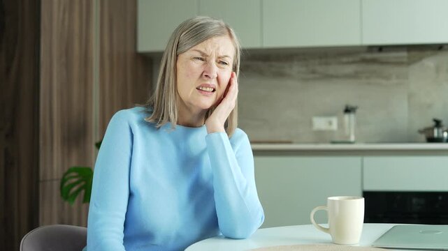 Sick senior female suffers from ear pain sitting in kitchen at home. Upset elderly woman touches her ear with her fingers. She has furuncle, shingles, eczema, ear diseases, otitis media. Close up.