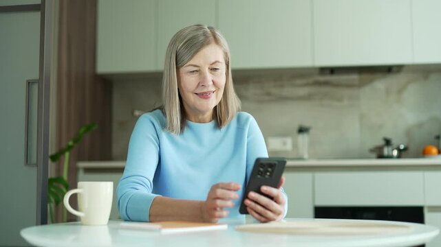 Smiling senior woman with gray hair using smartphone sitting in kitchen at home. Happy elderly female pensioner browses social media, chats online, writes or reads messages, shopping on mobile phone.