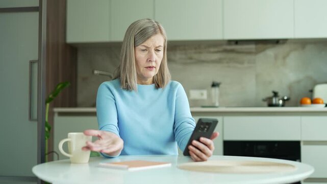 Confused senior woman having difficulty using smartphone sitting in kitchen at home. Worried aged female has app error, payment issue, received negative notification on phone and looking at camera.