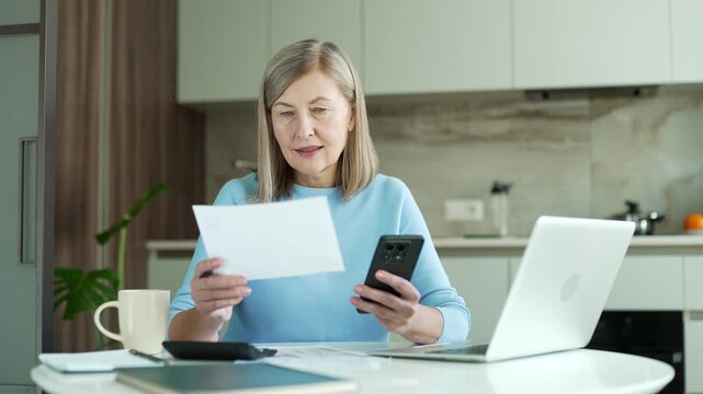 Happy senior female uses mobile app for spending bills on smartphone sitting at home office. Satisfied elderly woman calculates household finances, enters data into chat bot easy and simple to pay.