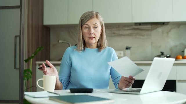 Confused elderly woman having difficulties with household finances documents using calculator while sitting in kitchen at home. Senior female dealing with problem in payments bills, unclear paperwork.