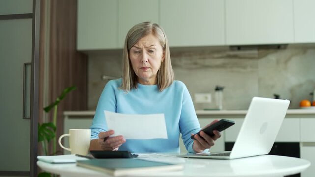 Confused senior female with gray hair having difficulty doing household finances using an application on mobile phone sitting in kitchen at home. Shocked elderly woman looks at large utility bills.