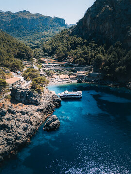 Aerial view of the azure sea meets the rugged coastline and verdant mountains where a boat rests gently near rocky outcrops, Mallorca, Balearic Islands, Spain.