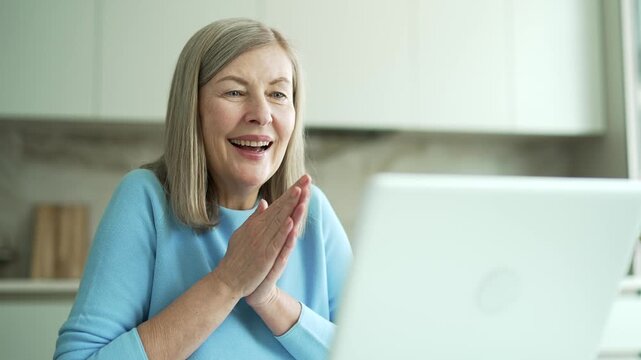 Happy excited senior woman with gray hair celebrating success by reading great news on laptop sitting in kitchen at home. Smiling elderly female satisfied with positive message on computer. Close up.