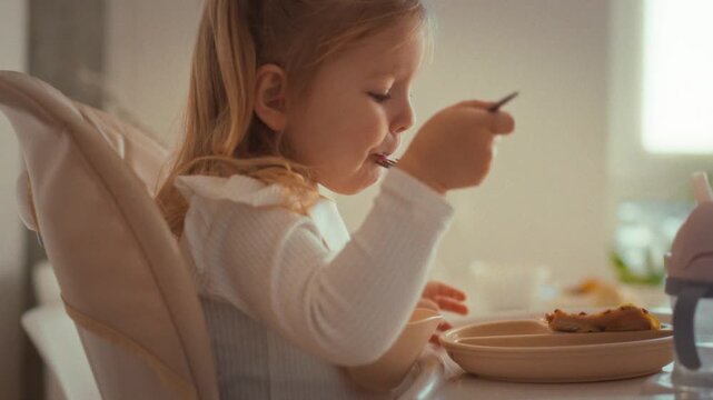 Seated cozy baby chair, little girl enjoys her meal independently, skillfully maneuvering fork while protected by colorful apron around her neck, embodying early childhood autonomy messy mealtime joy