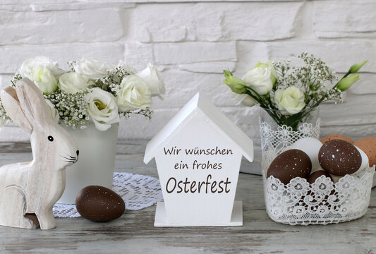 Deutscher Ostergru&szlig; &bdquo;Wir w&uuml;nschen ein frohes Osterfest&ldquo; mit wei&szlig;em Holzhasen, braunen Eiern und Fr&uuml;hlingsblumen vor einer wei&szlig;en Backsteinmauer.