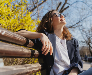 Obraz na płótnie Canvas Relaxed young curly woman in glasses enjoying spring sun on a city bench under blooming yellow forsythia bushes