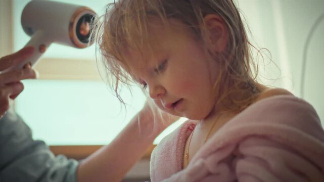 A young mother tenderly dries her little girl's hair with a hairdryer at home, moving slowly and cautiously to ensure the child's safety and comfort, showcasing a caring