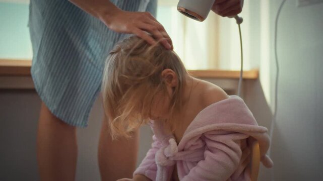 With tender care, young mother gently dries her baby's damp locks using a hair dryer, blowing soft, warm air to comfort her little daughter, ensuring a soothing and nurturing experience for the child