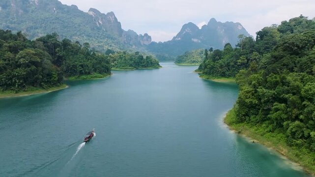 Aerial view of a longtail boat slicing through the turquoise waters of Cheow Lan Lake, surrounded by lush green rainforest and towering limestone cliffs, Khao Sok, Thailand.