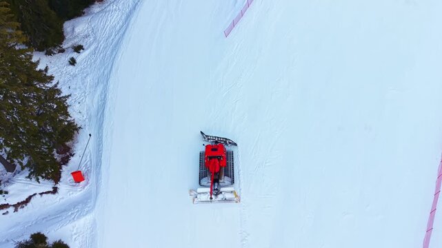 A snow groomer works on a ski slope in the mountains at a winter resort. The machine clears and smooths the snow for skiers. Tall trees surround the area. Bright snow covers the ground.