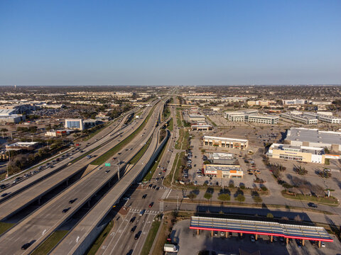 Aerial view of a highway cutting through the urban landscape, lined with buildings and gas stations under a clear sky, Frisco, Texas, United States.