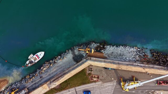 Workers use machinery to add rocks along the coast while a boat is anchored nearby in a port area during the day.