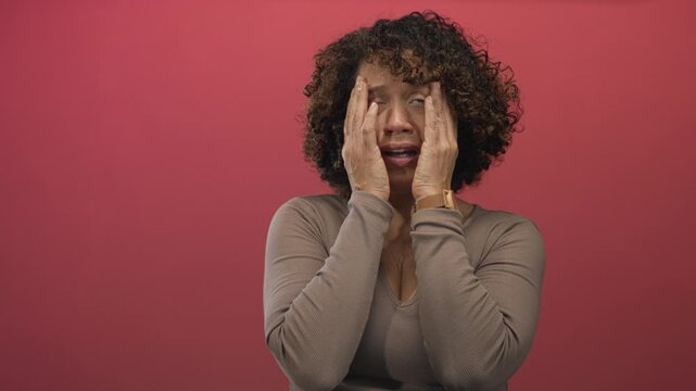 Hispanic middleaged woman holds face with hands in pink studio setting displaying concern; emotional distress.