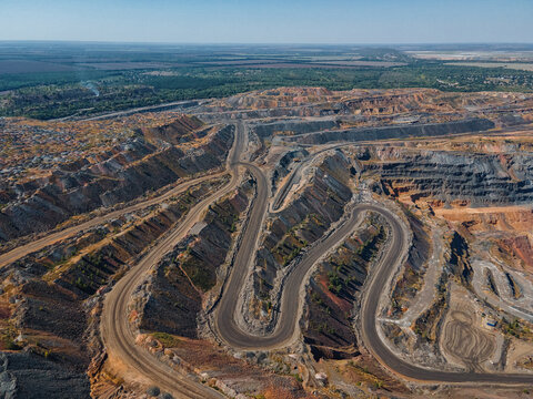 Aerial view of a winding road cuts through the stark, layered landscape of the vast iron ore quarry, contrasting with the distant green forests, Kryvyi Rih, Dnipropetrovsk Oblast, Ukraine.
