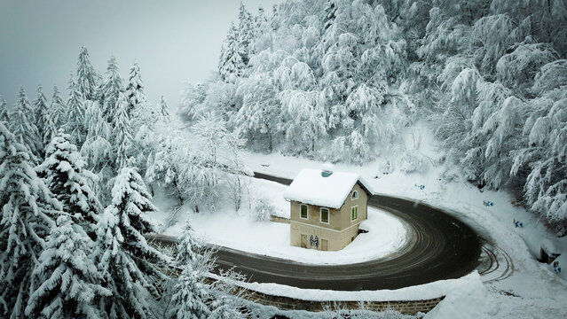 Aerial view of a solitary house on Col de la Faucille road surrounded by snow-laden trees and a winding road, nestled in a winter wonderland, Gex, Auvergne-Rhone-Alpes, France.