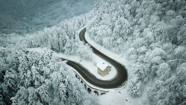 Aerial view of a lone house nestled in a snowy curve on Col de la Faucille road, surrounded by a forest blanketed in white, Gex, Auvergne-Rhone-Alpes, France.