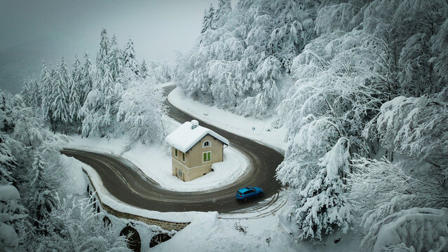 Aerial view of a blue car navigating on the road to Col de la Faucille past a solitary house amidst a forest blanketed in white, Gex, Auvergne-Rhone-Alpes, France.