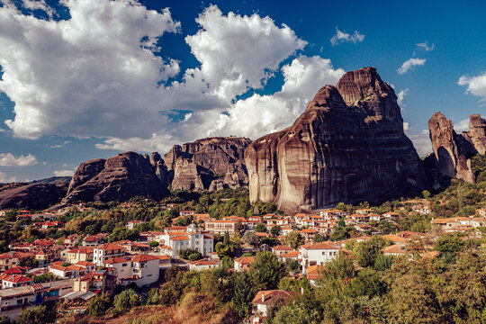 View of ancient rock formations tower over a quaint village with red-tiled roofs, nestled amidst lush greenery under a sky of swirling clouds, Kalabaka, Trikala, Greece.