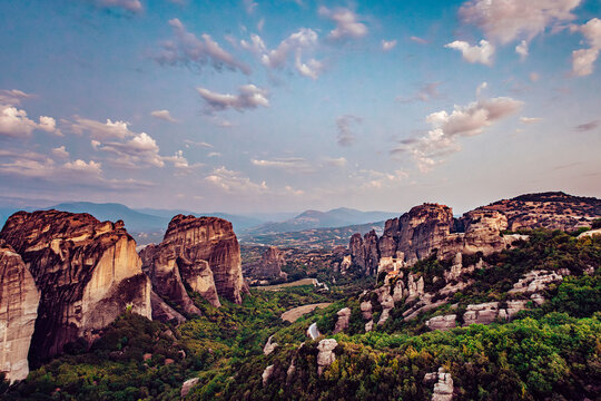 View of towering rock formations, green vegetation, and distant mountains under a bright sky, bathed in golden light, Kalabaka, Trikala, Greece.